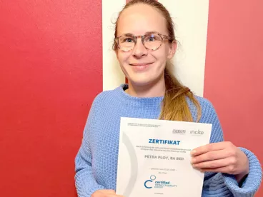 Petra Ploy ist Certified WebAccessibility Expertin Eine Person hält eine Urkunde vor einem rot-weißen Hintergrund und lächelt in die Kamera.